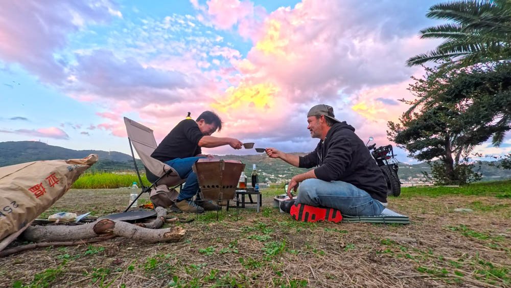 Two men sharing drinks beside a campfire at sunset during a motorcycle trip in Ehime