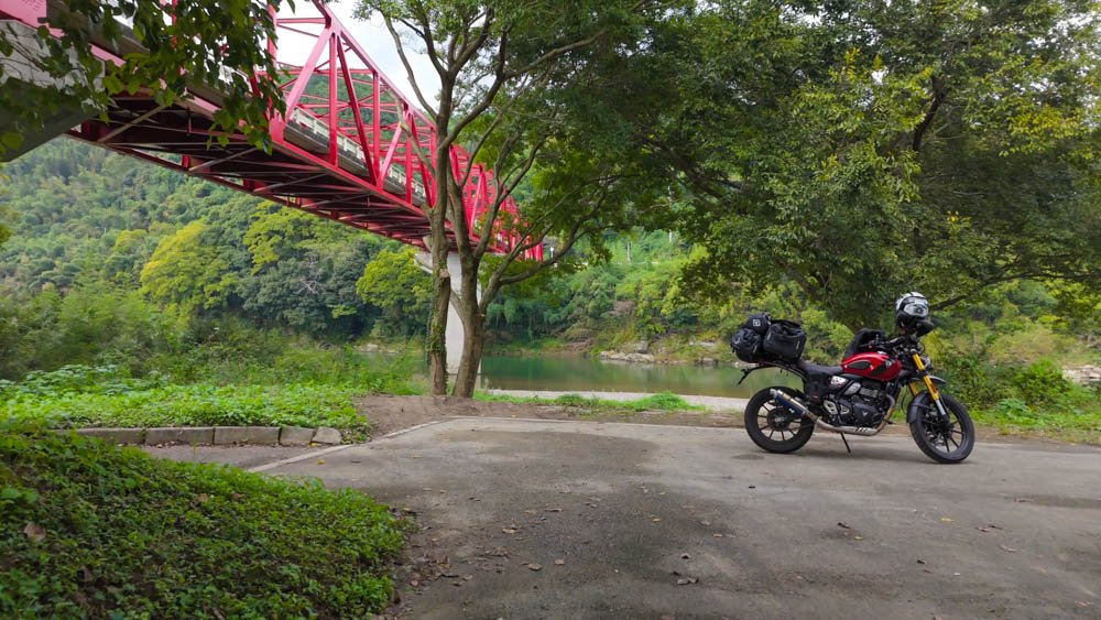 Red Triumph Scrambler parked beneath a red bridge beside a river in Ehime