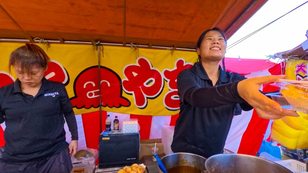 Smiling food vendor serving takoyaki at a local festival in Matsuyama, Ehime