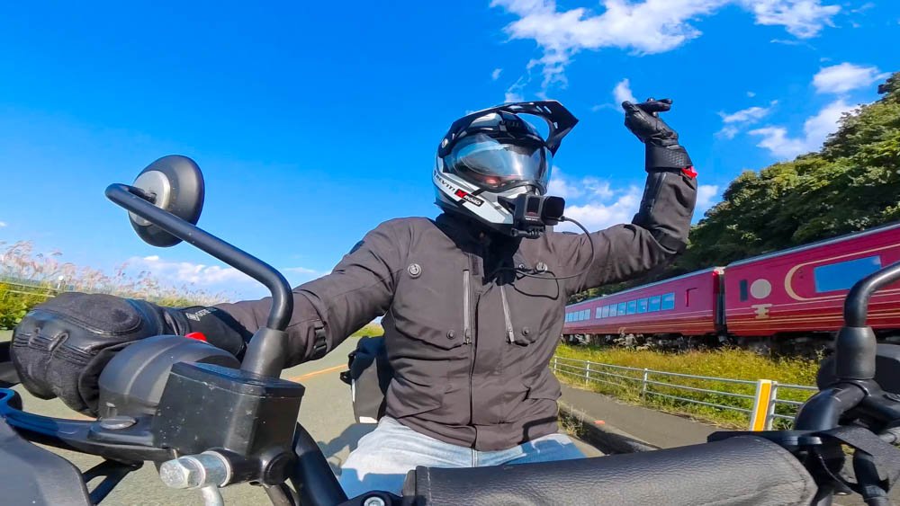 Helmet-camera view of a motorcycle rider beside a red passenger train on the Ehime coast