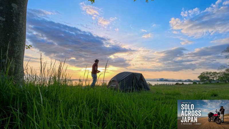 Solo motorcycle traveller standing beside a tent at sunset on Lake Biwa